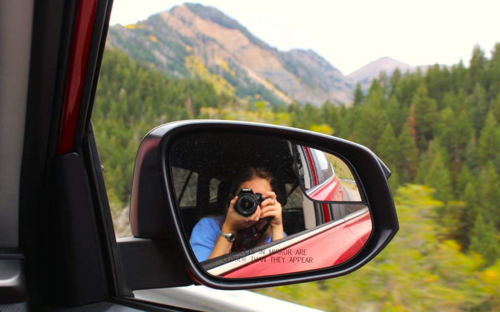Photograph of Brianna Eisman taking a photo in the mirror of a car driving in the Utah mountains in the Alpine Loop